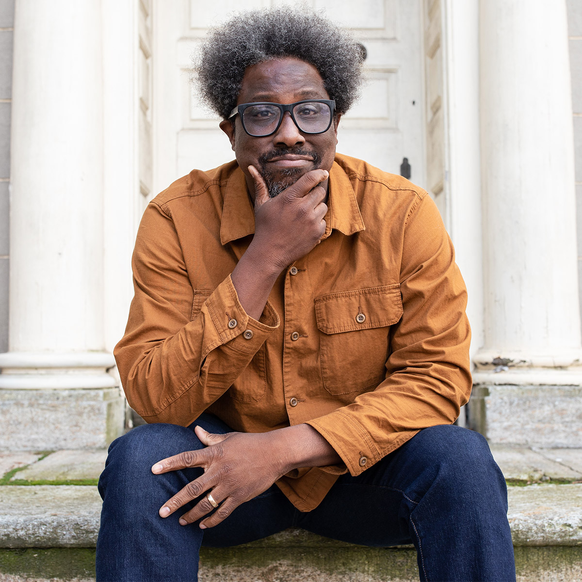 W. Kamau Bell is a Black man seated on the stone steps of a building that we can only see the bottom of. He has one hand resting open on his knee and another bent at the elbow resting on the same knee and his hand resting on his chin and he softly smiles.
