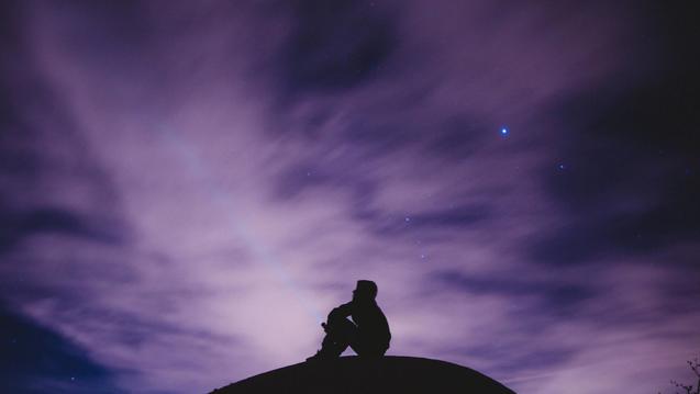 Photo of a silhouetted person sitting atop a mound with a starry purple sky in the background.
