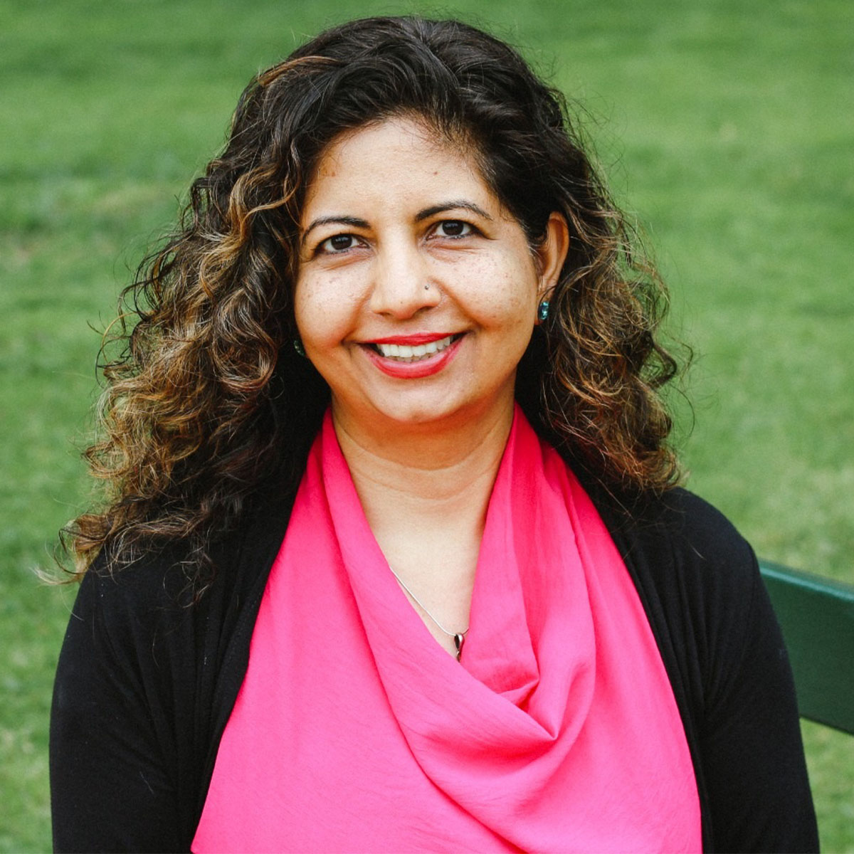 Alka Arora color portrait. Alka is an Indian American woman with dark brown curly hair. She is smiling and seated on a bench outside with grass behind her.