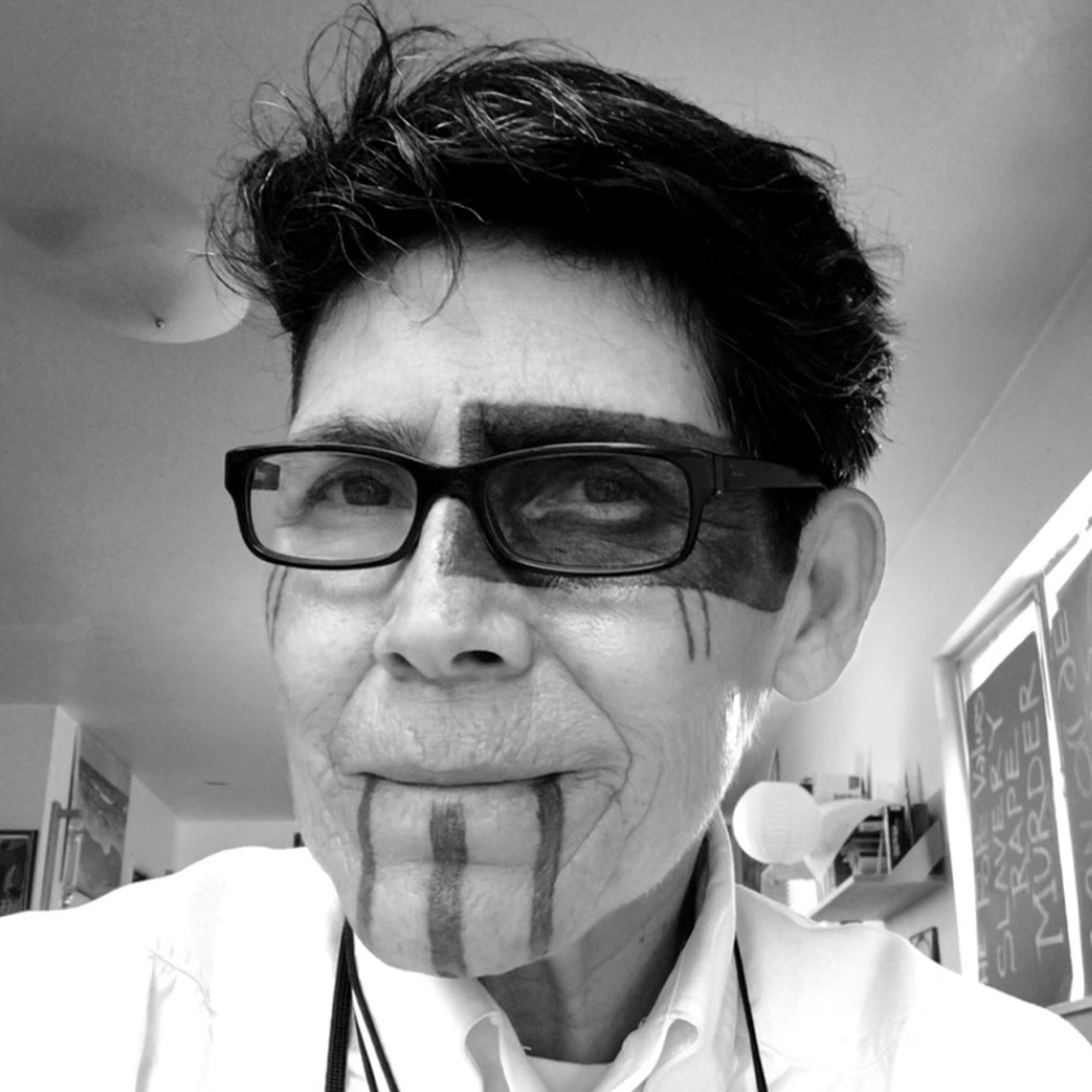L. Frank black and white portrait. L. Frank is Tongva & Ajachemem & Rarámuri Two-Spirit person. Her hair is short and is a dark-color. In the photo L. Frank is wearing dark-rimmed glasses, and has Indigenous tattoos over one eye and three lines down her chin.