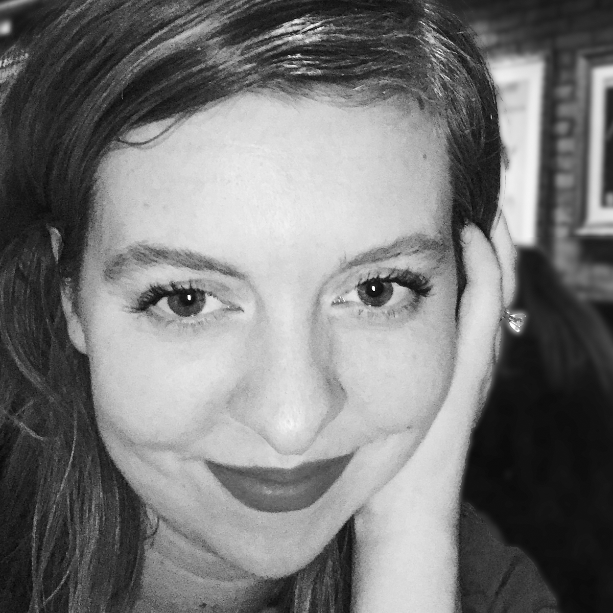 Larisa Garski black and white portrait. Larisa is posed close to the camera and her full face fills the shot. She is smiling, has dark-colored hair pushed behind her ears. She has one hand resting on the side of her head.