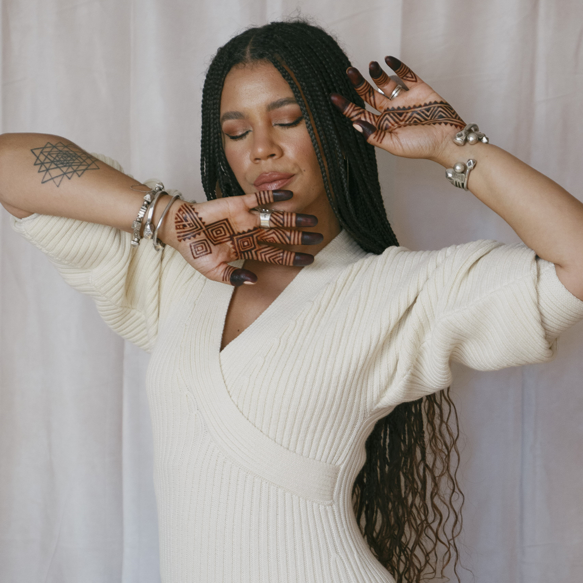 Maryam Hasnaa color portrait. Maryam is a Black woman with long, braided hair to her lower back. Her eyes are closed and her arms are raised and hands open across her chin and head.