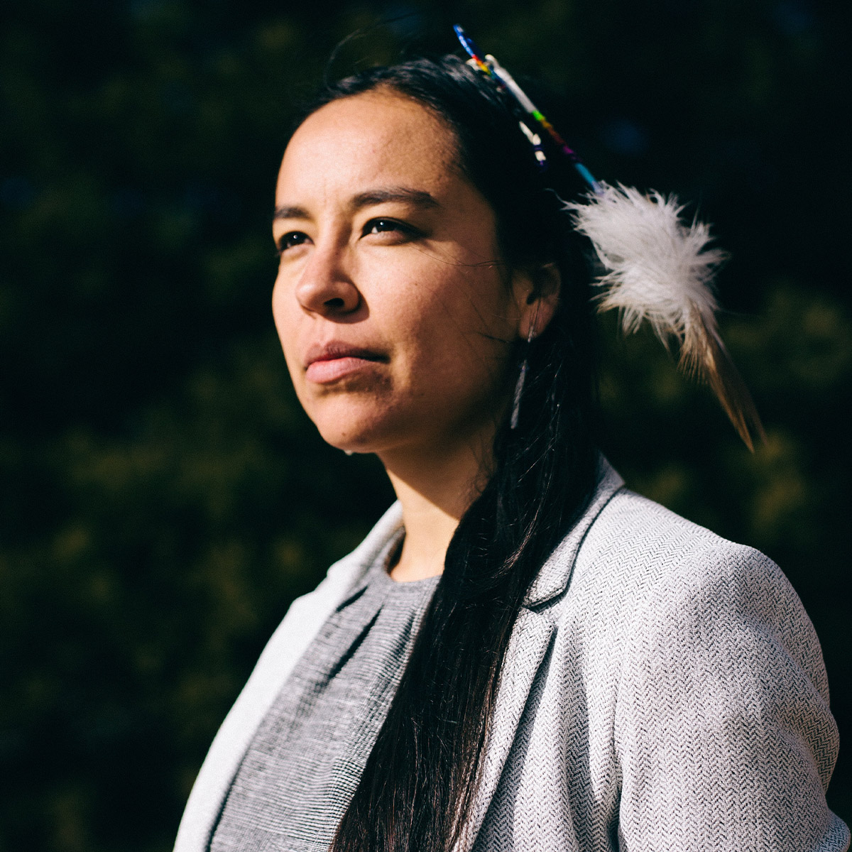 Lyla June color portrait. Lyla is Diné (Navajo), Tsétsêhéstâhese (Cheyenne), and European lineages. She is looking off to the far left and is in partial profile. She is wearing beads and feathers from her cultural communities in her long, dark-colored hair. She is also wearing light gray colored blazer and top. Behind her appears to be greenery so this photo may have been taken outside.