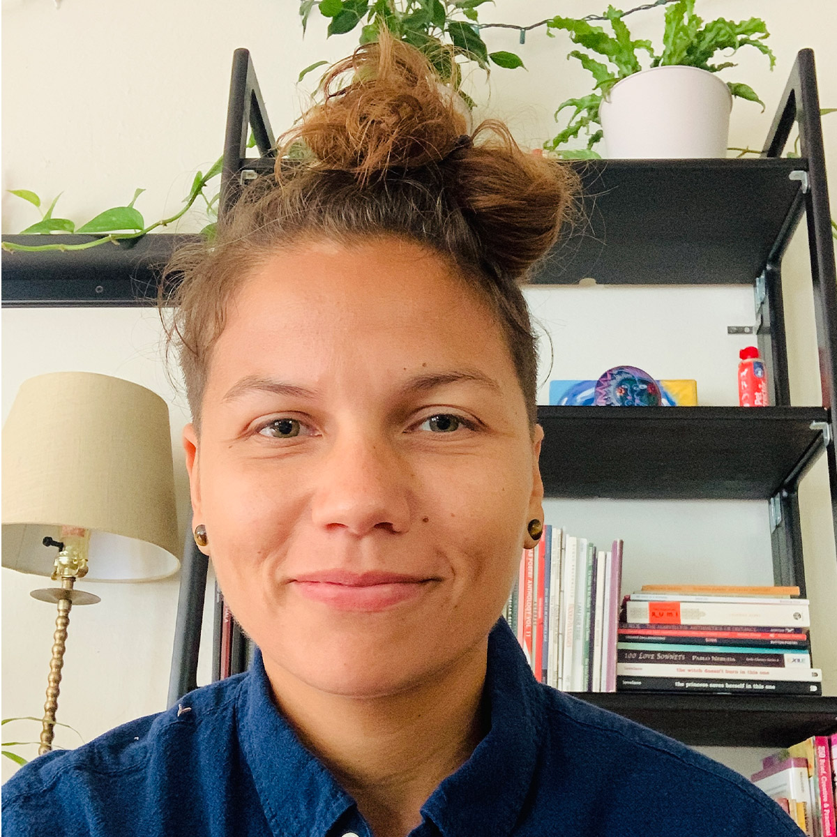 Jenna Robinson color portrait. Jenna is a Queer Woman of Color and is posed in from of her bookshelf filled with books and plants. She is smiling and has her brown hair tied into a bun on top of her head.