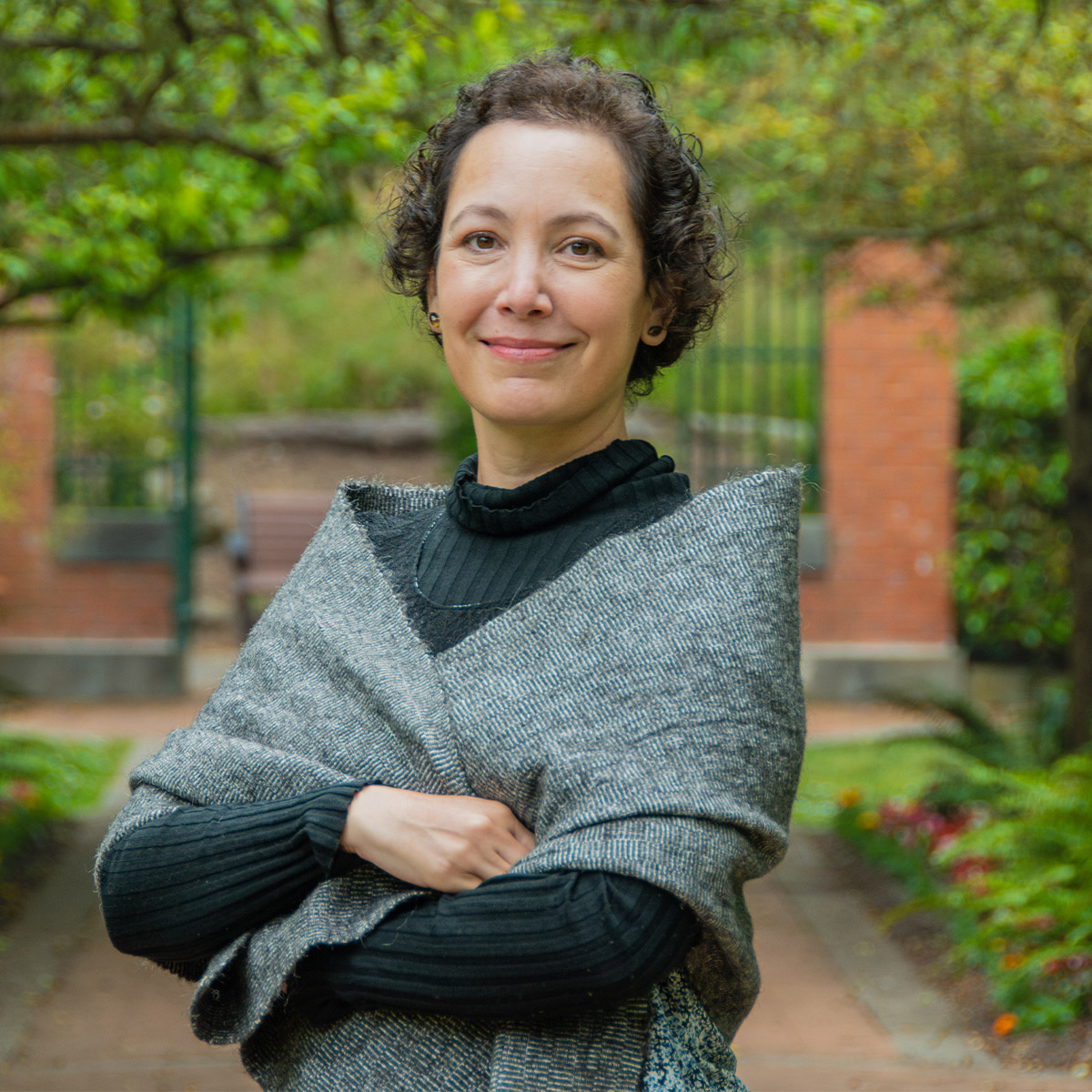 Charlotte Seanz color portrait. Charlotte is posed with arms crossed in front of a brick and metal gate. She is smiling and wearing a dark-colored turtleneck and gray shawl.
