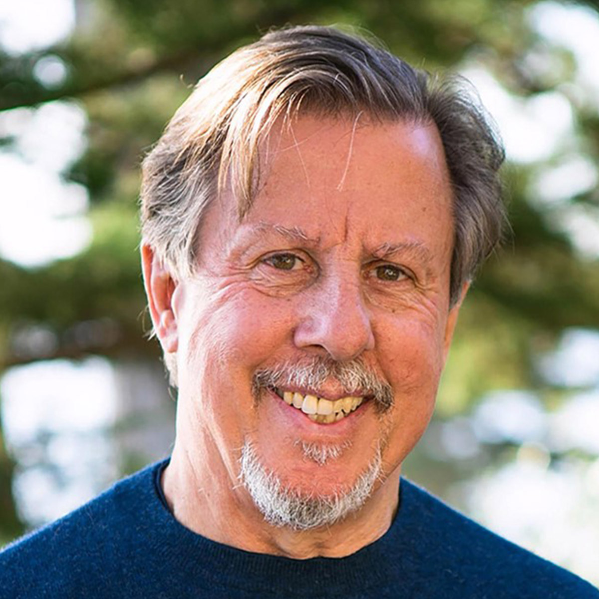 Richard Schwartz color portrait. Richard is an older, white man with silver, brown hair that is short. He is smiling and is posed outside with a tree blurred in the background.