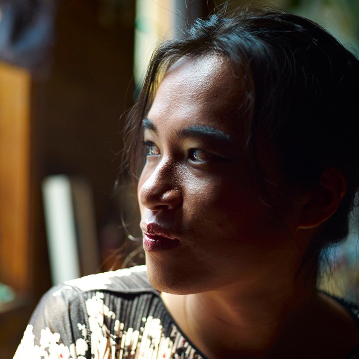 Kai Cheng Thom color portrait. Kai Cheng is in part profile and is looking off to the left, to what appears to be a light source, maybe a window. They are wearing a silk top with pleats and gold design.