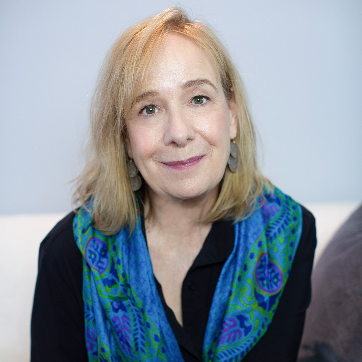 Suzi Tucker color portrait. Suzi is an older, white woman with medium-length blonde hair and is smiling. Suzi is seated and is earing a bright green and blue printed scarf and a dark-colored top.