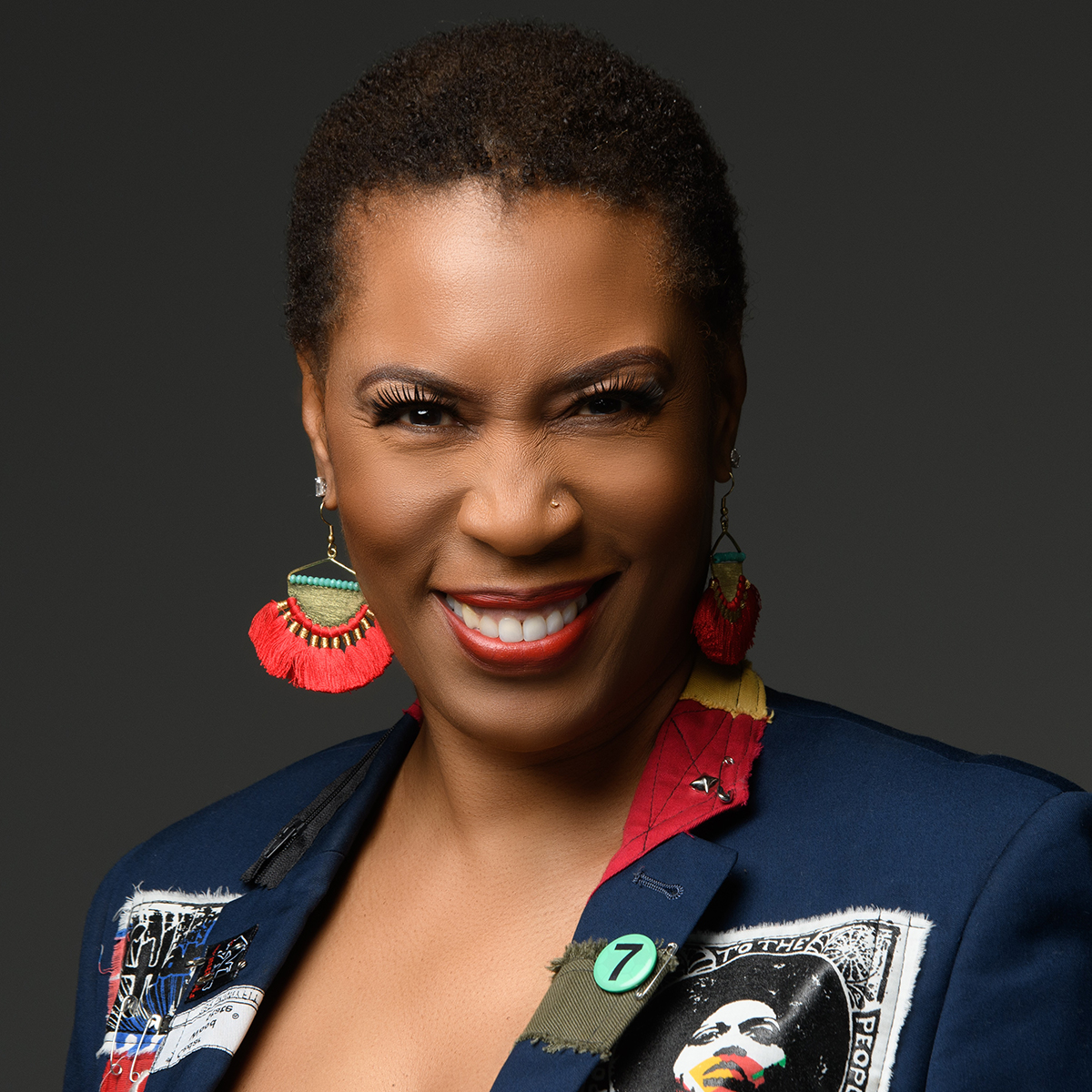 Jenée Johnson color portrait. Jenée is a Black woman with short hair, bright red lips, and brightly colored, dangling earrings. She is wearing a blue blazer with patches, textures, different fabrics across it.