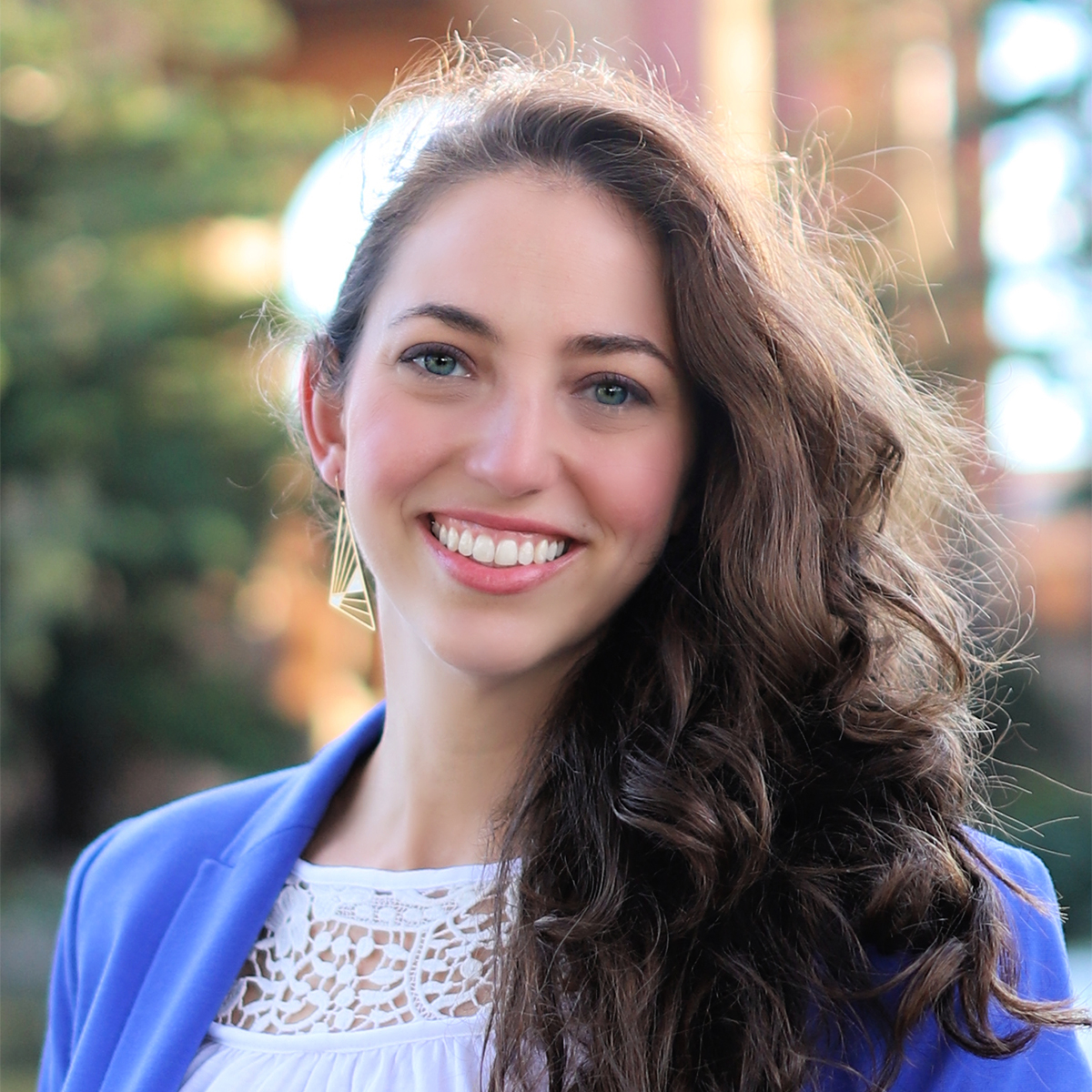 Laura Pustarfi color portrait. Laura is smiling and posed outside. Her hair is wavy, curled and is over one shoulder. She is wearing a blue blazer and white top.