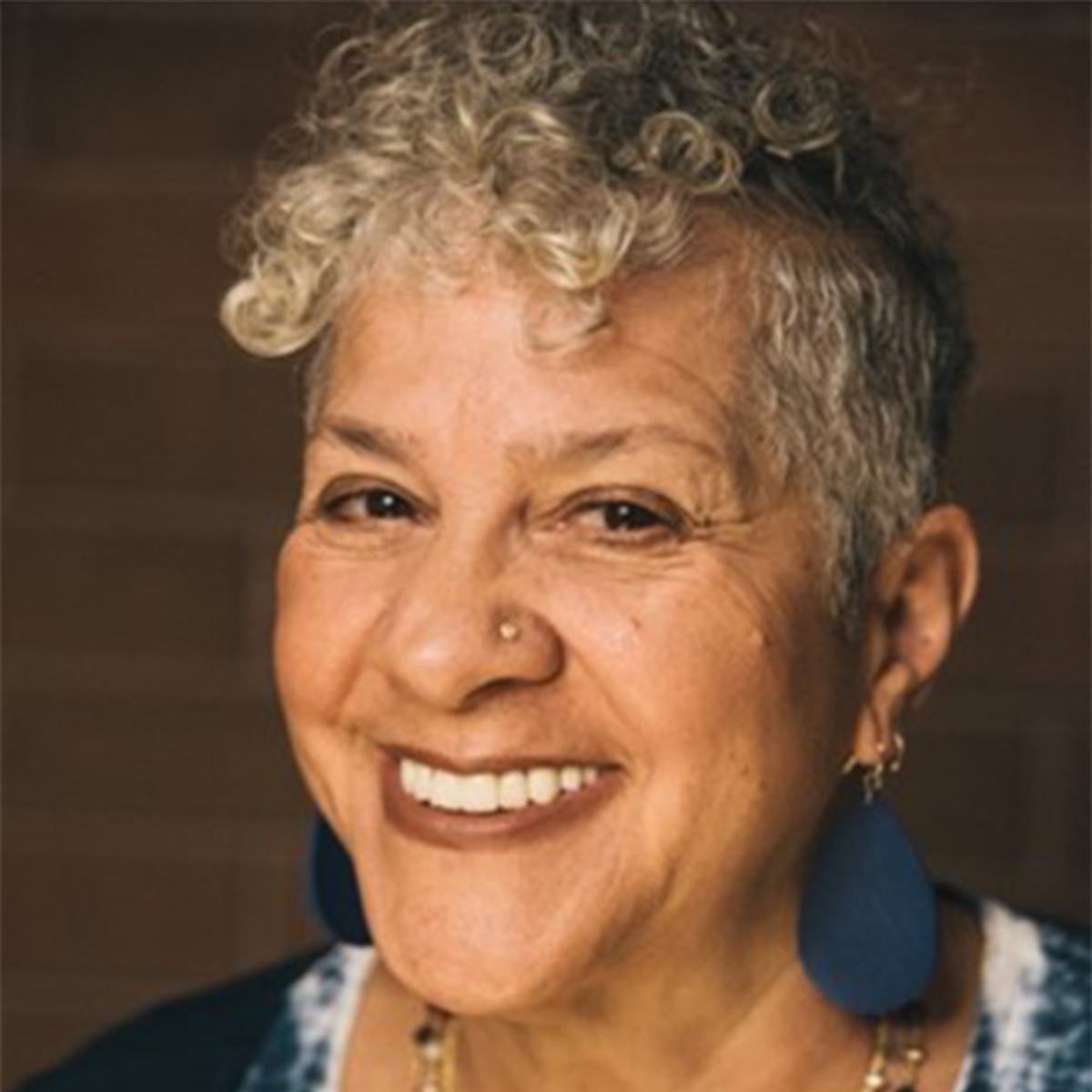 Shakti Butler color portrait. Shakti is a Black, older woman with short, hair that is curly, light in color, and shaved around the temple and around the middle of her head. She is smiling and wearing blue earrings and has a small stud nose piercing.