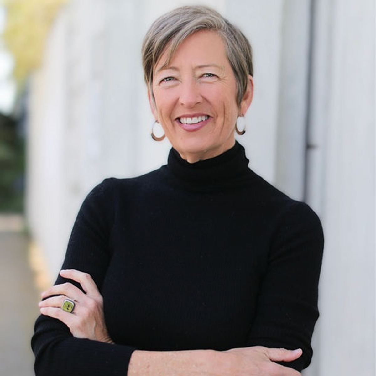 Staci Haines color portrait. Staci is a white woman with short brown, gray hair. Staci is smiling and wears gold hoop earrings and a black turtleneck and her arms are crossed.