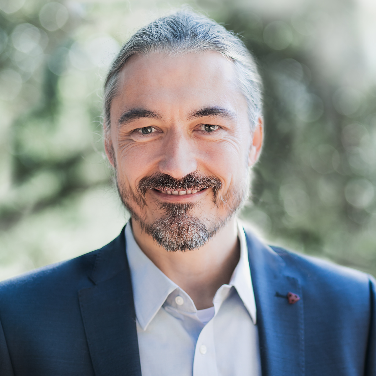 Thomas Hübl color portrait. Thomas is smiling, posted outside, and is wearing a blue blazer, and light-colored button down. He is an older white man with his silver, gray hair pulled back and his a light beard.