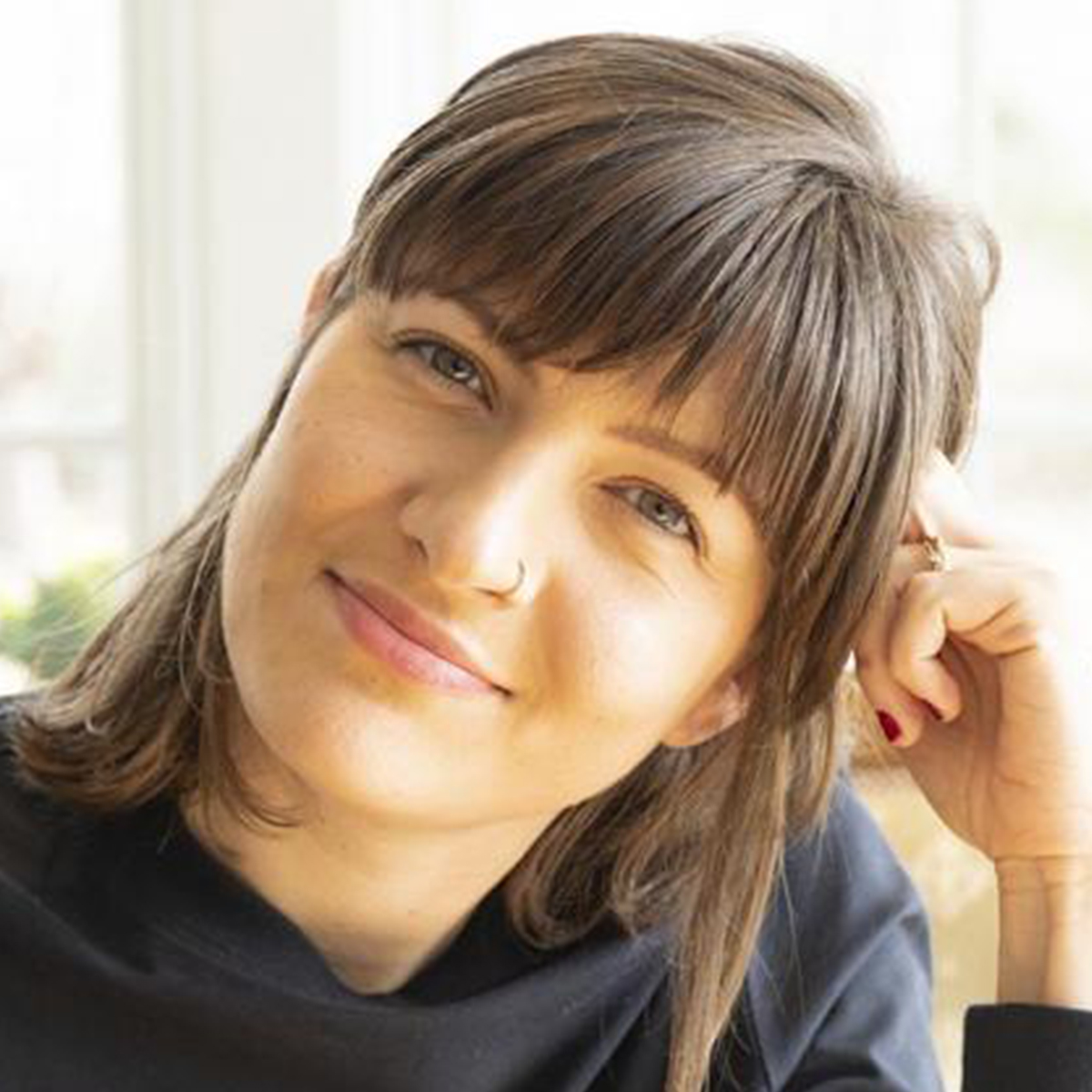 Mirjam Paninski color portrait. Mirjam is a white woman with brown hair that ends at her shoulders. She is smiling and has her head tilted and rested on her right hand.