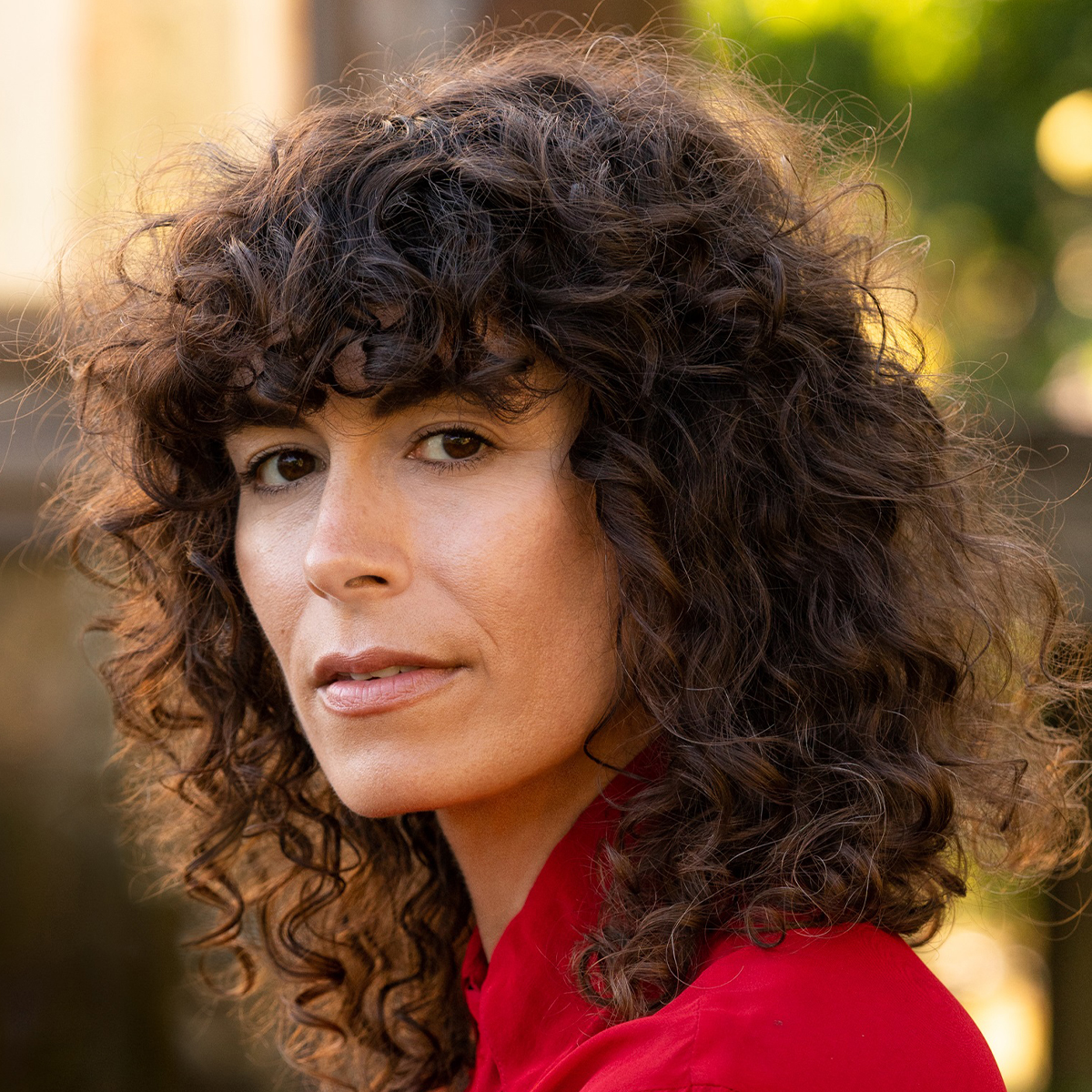Jacqueline Suskin color portrait. Jacqueline has her head turns towards us (or the camera) and is looking straight ahead. Her mouth is slightly open and she appears to be outside. Her hair is dark brown and curly and is wearing a deep red top.