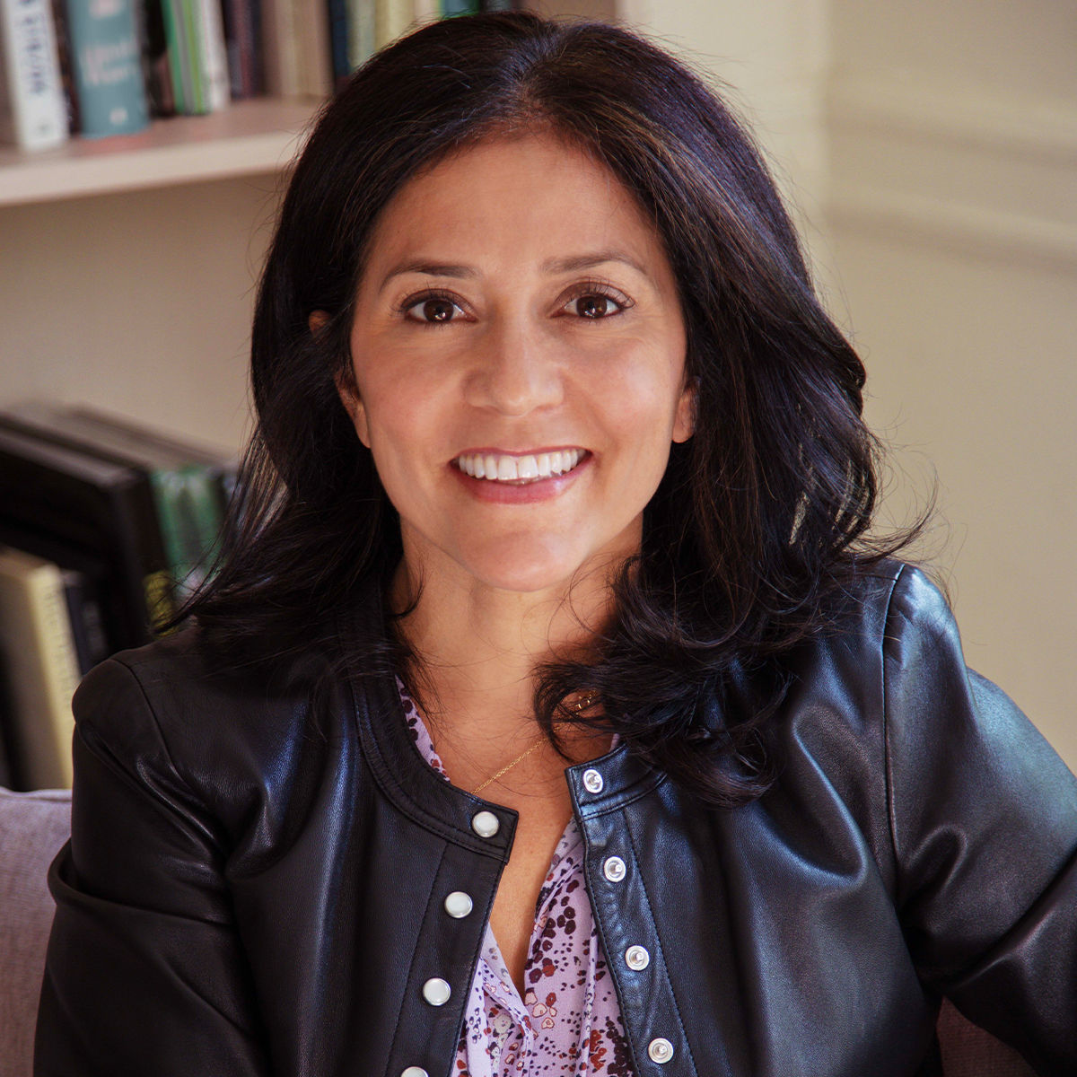 Lisa Weinert color portrait. Lisa is seated and is wearing a black leather jacket and floral top. She is smiling and has dark brown hair that ends just below her shoulders.