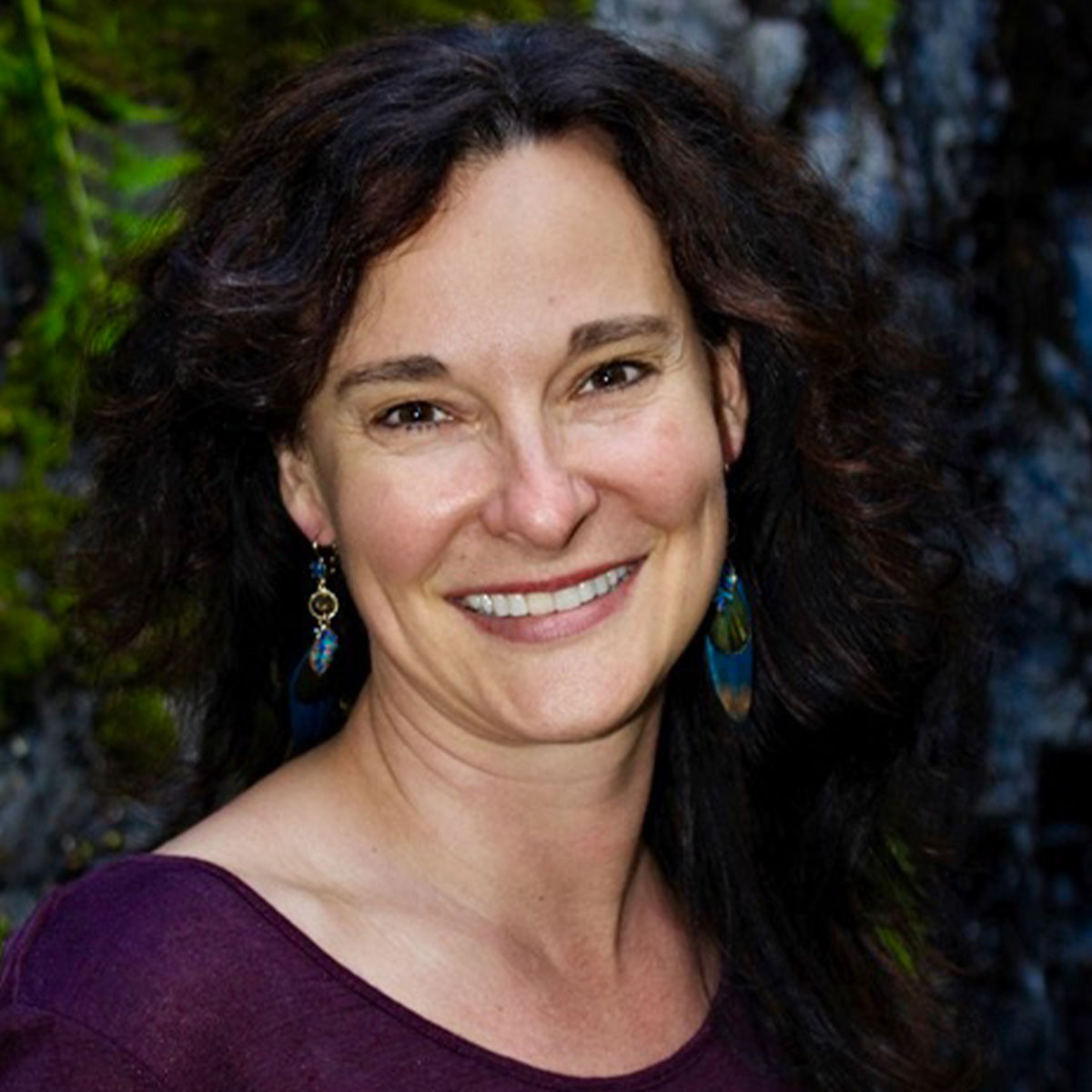 Prema McKeever color portrait. Prema is a white woman with dark brown curly hair that ends after her shoulder blades. She is smiling, wears dangling blue earrings and a deep purple top.