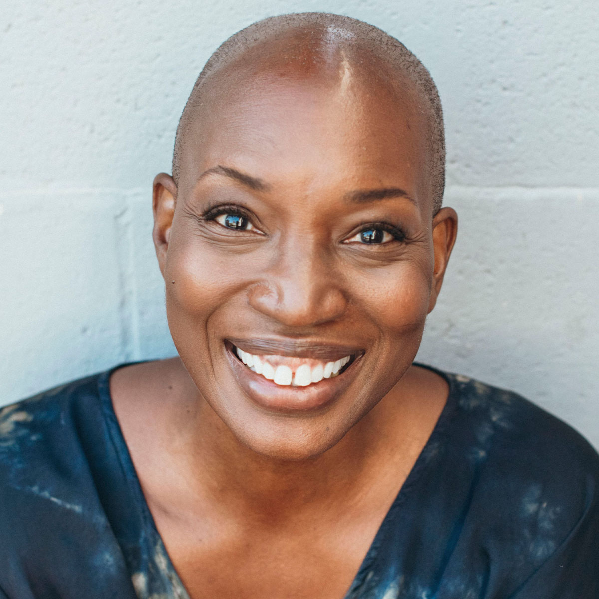 Tracee Stanley color portrait. Tracee is a Black woman leaning against a light-colored wall. Tracee is smiling and wearing a subtly printed top.