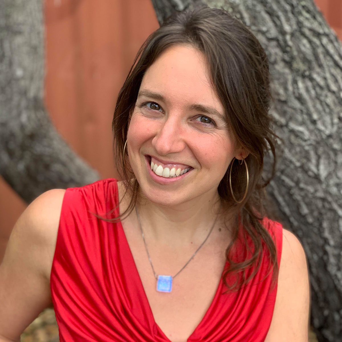 Marie Thouin color portrait. Marie is a white woman with brown hair pulled back with strands falling around her shoulders. She is smiling, wearing a red top, and a bright blue pendant as she poses by a tree which you can see the branches of behind her.