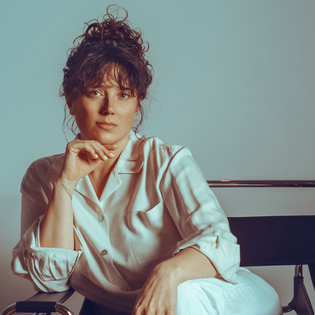 Abigail Rose Clarke color portrait. Abigail has brown, wavy hair, pulled up in a bun. She is seated and wearing a white button-down with sleeves rolled up at mid-length. One arm is bent, elbow on the armrest, and her hand is resting on her chin.