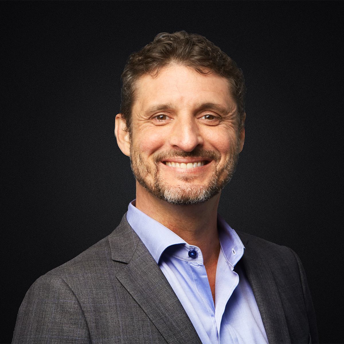 Drew Dillinger color portrait. Drew is a white man with short brown hair and a light beard. He is smiling, wearing a gray suit, and is posed in front of a black background.