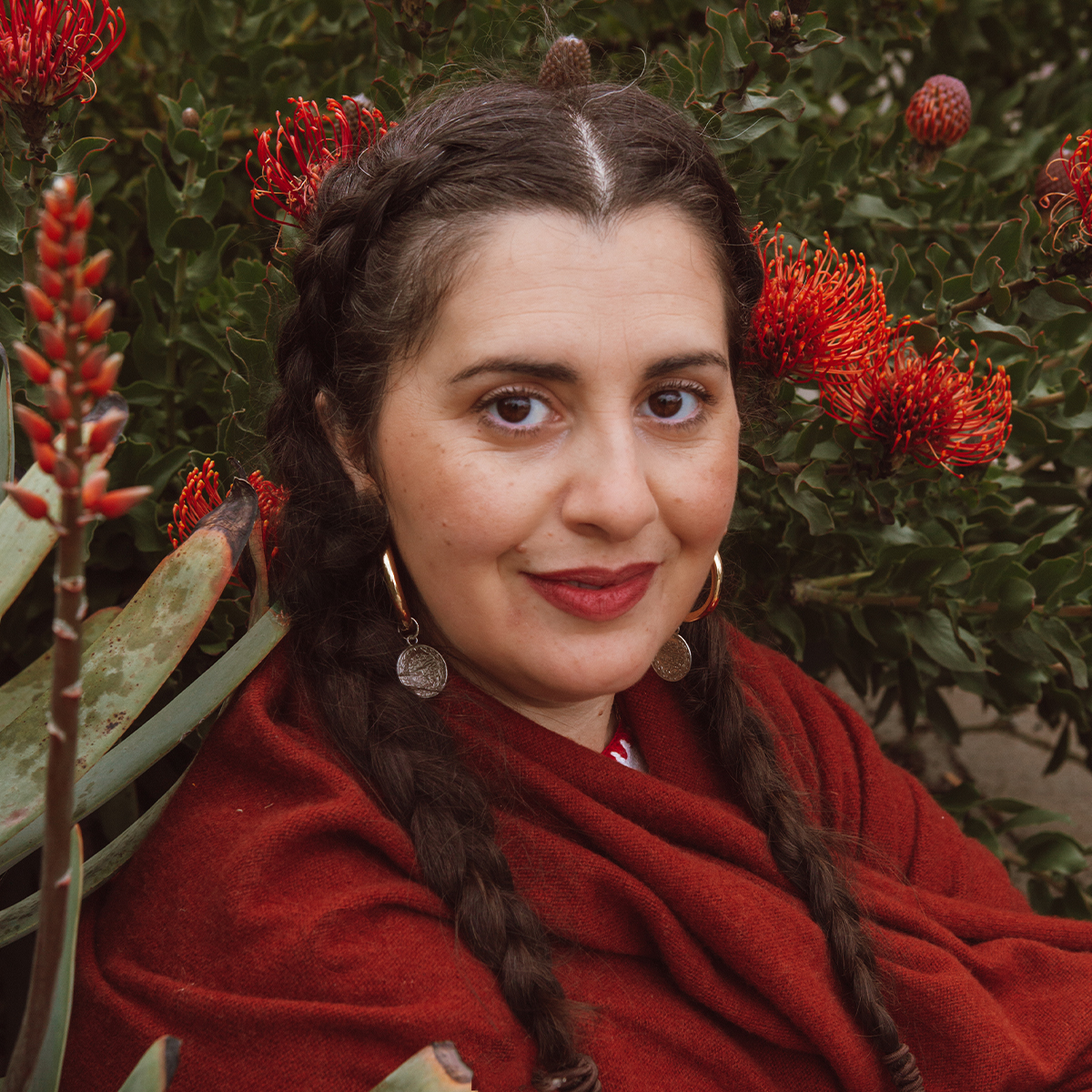 Layla K. Feghali color portrait. Layla is Lebanese American and has long dark brown hair that is braided and goes down past her shoulders. She is smiling, wearing a red shawl, and is posed outside amongst red flowers.  