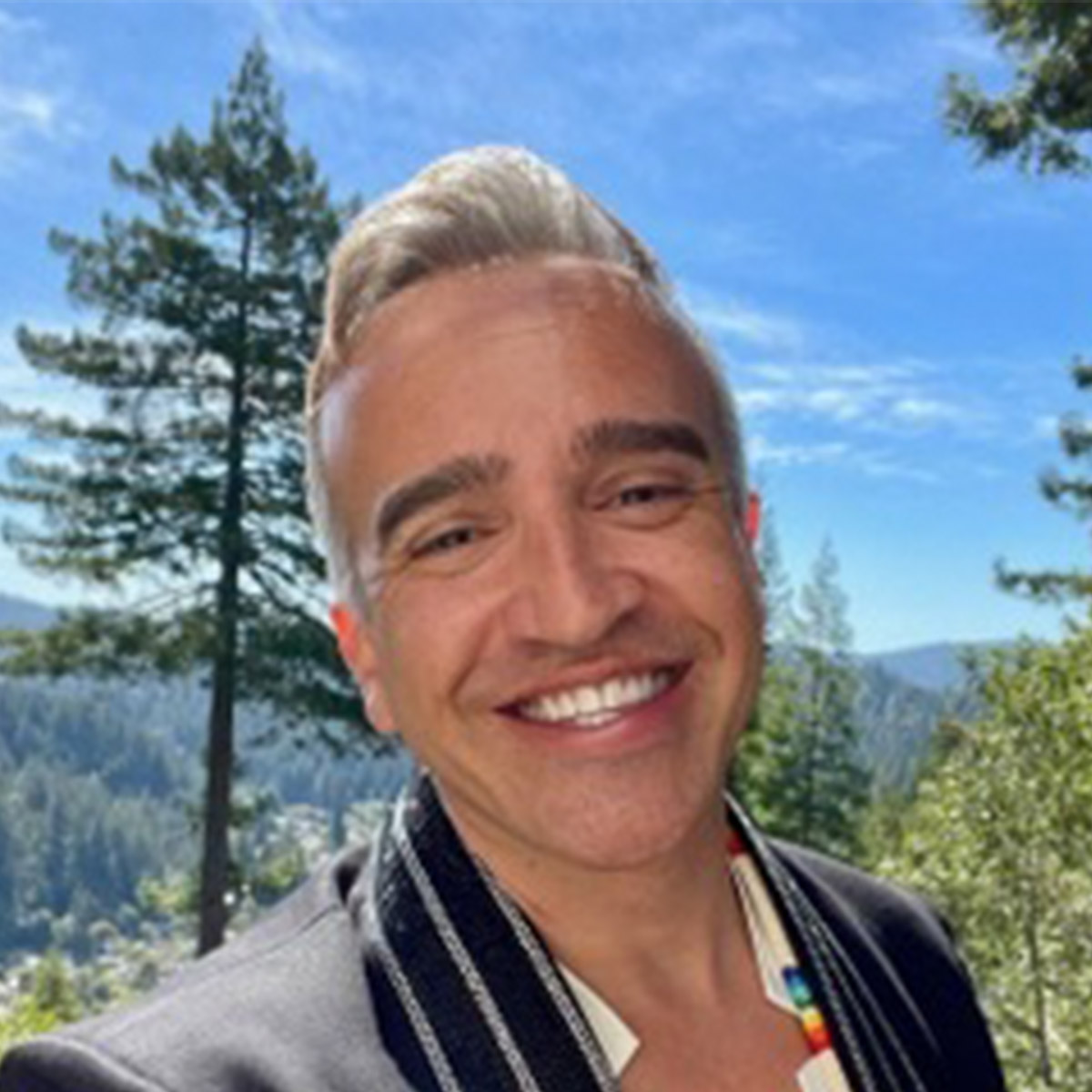 Roger Kuhn color portrait. Roger is a Poarch Creek Two-Spirit Indigequeer and is posed outside among pine trees and a blue sky. Roger is smiling, wears a dark-colored blazer.
