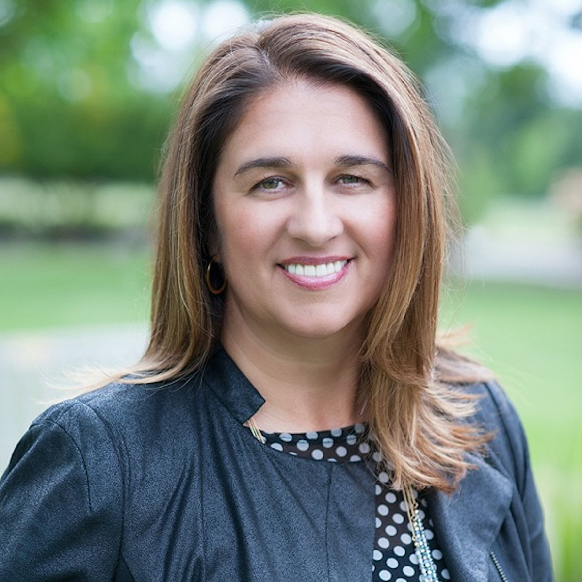 Manuela Mishke-Reeds color portrait. Manuela is of German and Spanish descent and has light-colored skilled, brown, straight hair that ends past her shoulders. She is smiling and wearing a dark-gray moto-style jacket and a polka-dotted top.