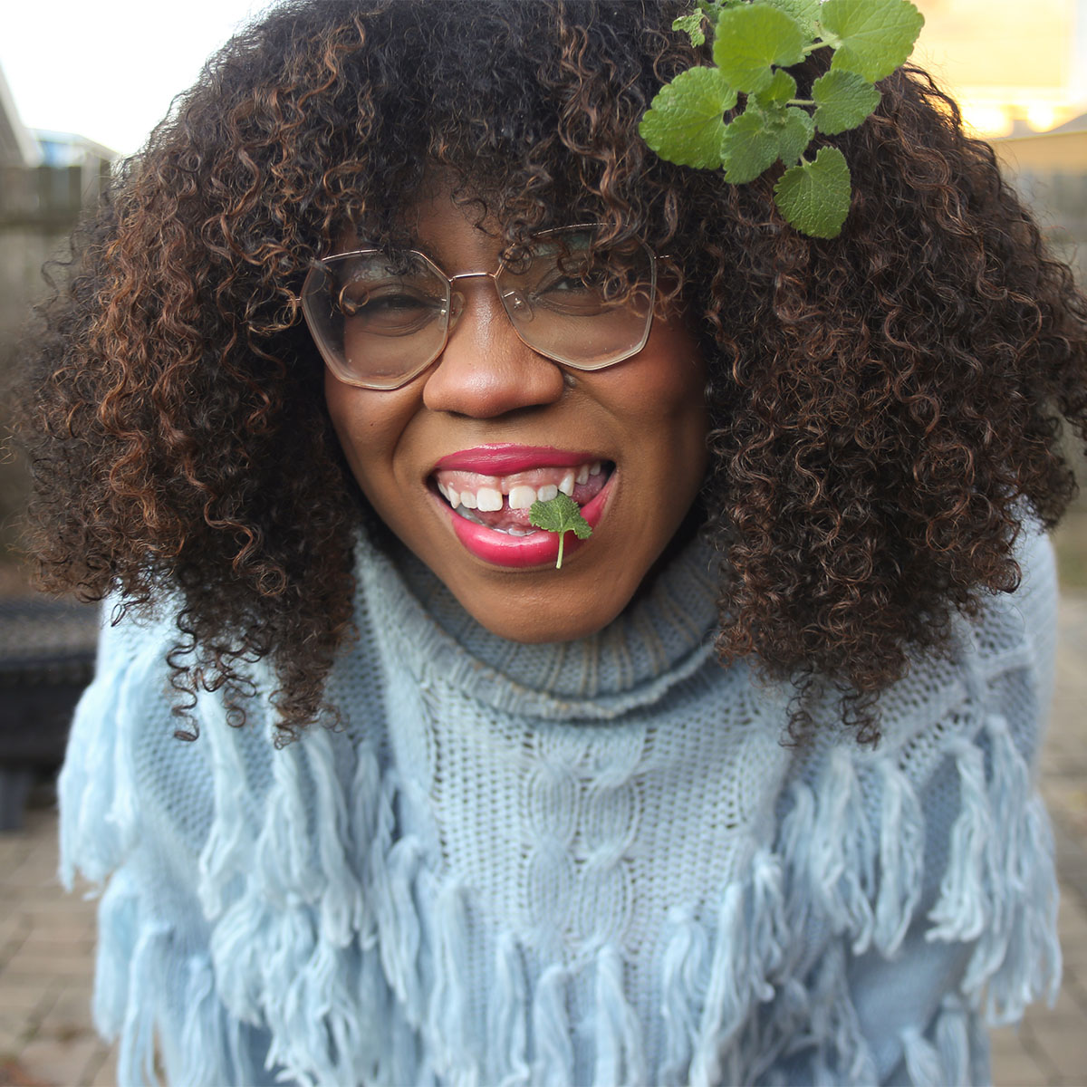 Alexis Nikole Nelson color portrait. Alexis aka Black Forager is a Black woman with shades of brown curly hair. She is wearing large glasses, red lips, and a blue knit sweater. She also has what looks like mint in her hair and is biting a bit of a green plant between her teeth as she smiles.