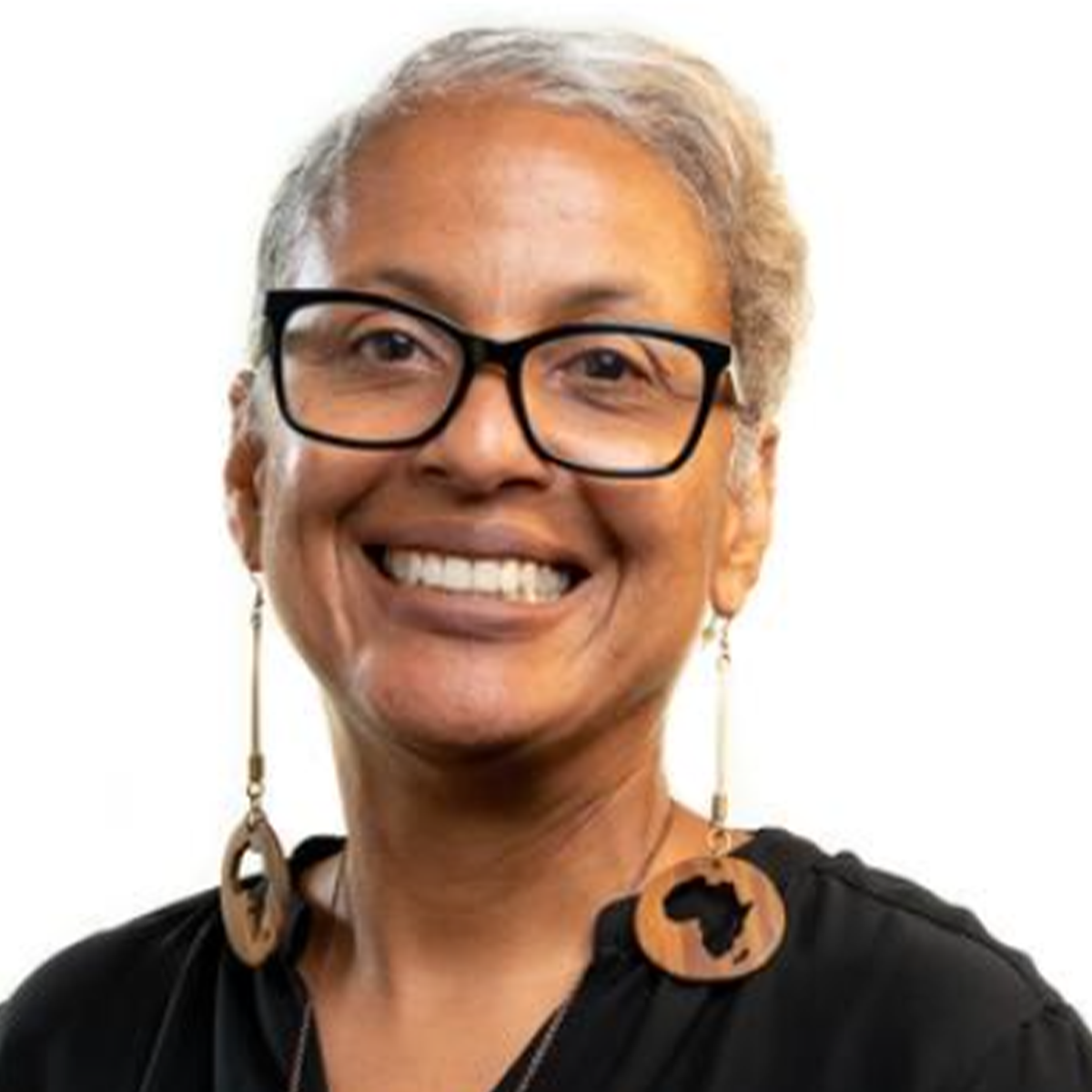 Damali Robertson color portrait. Damali is a Black-bodied woman of Caribbean and American descent; Jamaican by lineage, American by birth. She has short silvery hair, long wooden earrings with the African continent cut out at the center. She is wearing a black top, glasses, and is posed in front of a white background.