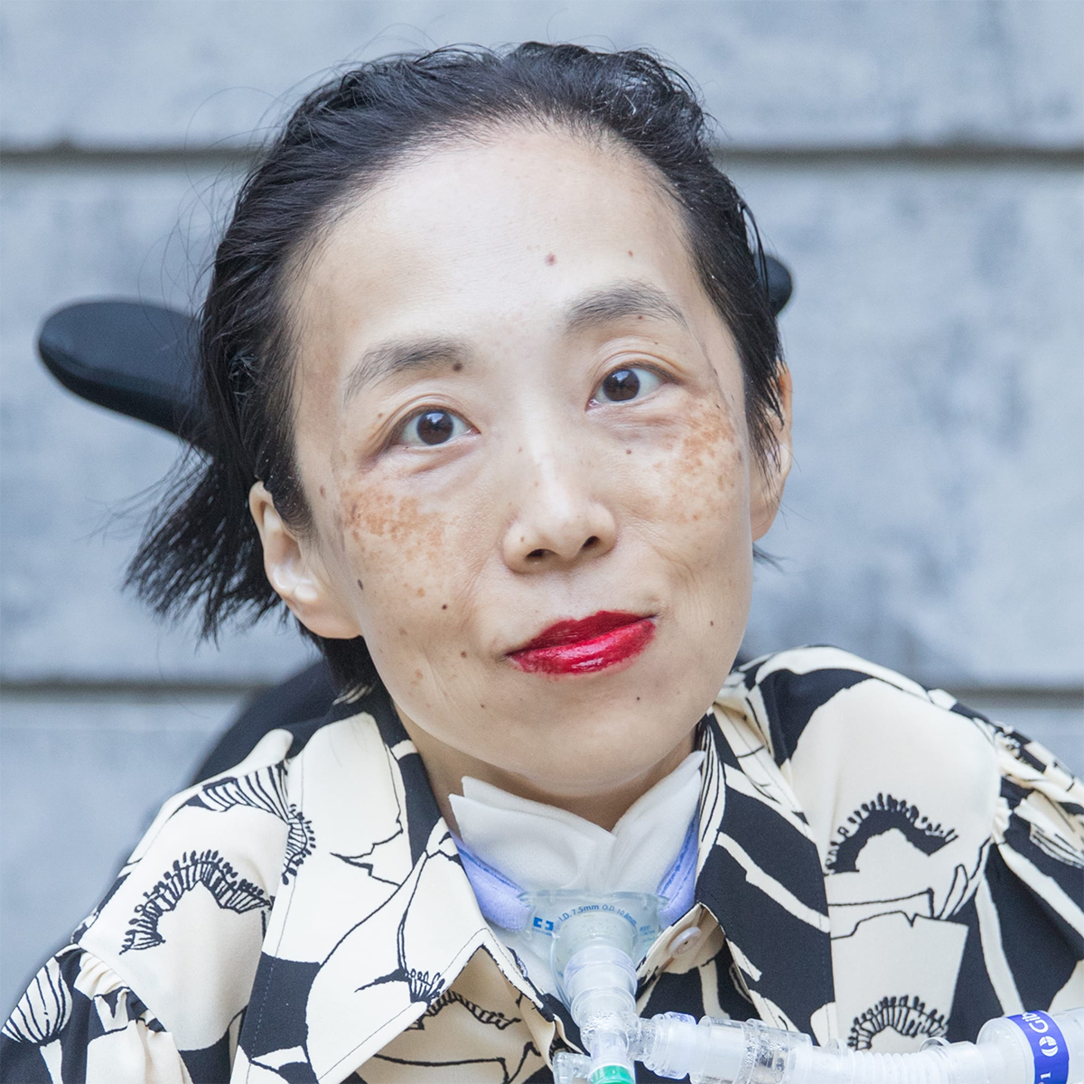 Alice Wong color portrait. Alice is Asian American disabled woman in a wheelchair with a tracheotomy with a tube attached to a ventilator. Alice is wearing a black and white floral patterned top.