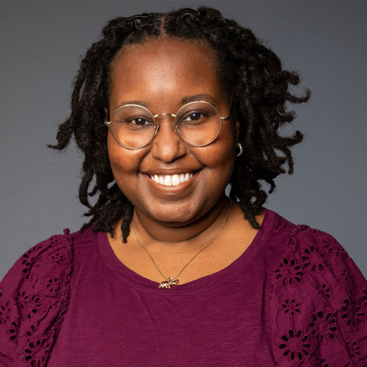 Britton Williams color portrait. Britton is a Black woman who is smiling in this portrait. Britton is wearing round, thin-framed glasses and a bright purple, and a pendant necklace.