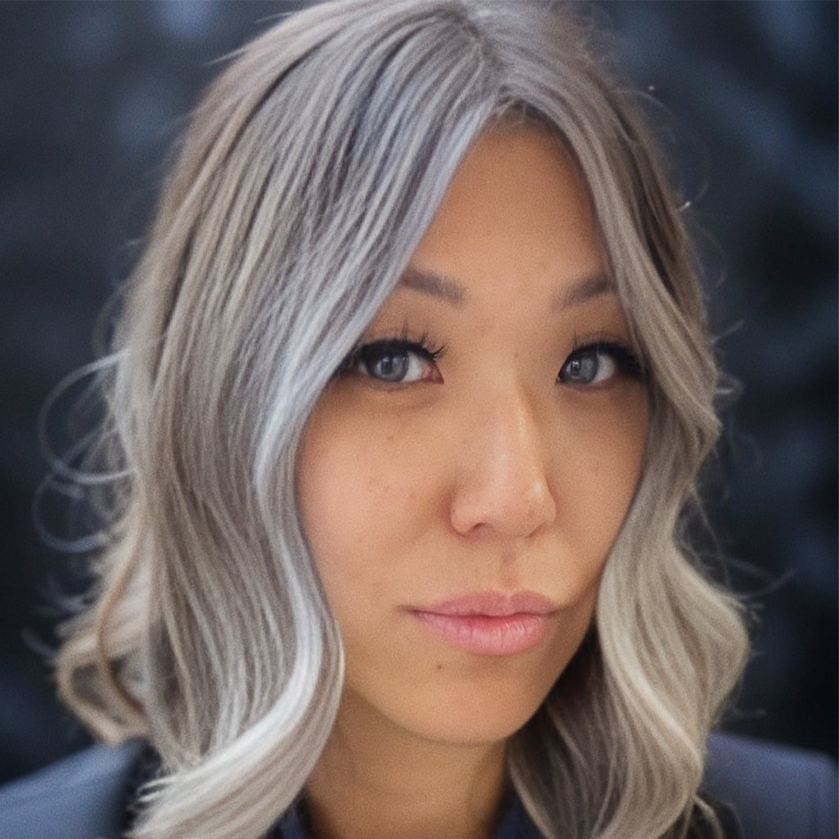 Kini Chang color portrait Kini is an Asian woman with wavy, medium-length hair in shades of light brown, light blond, silver, gray. She is posed in front of a blurred background