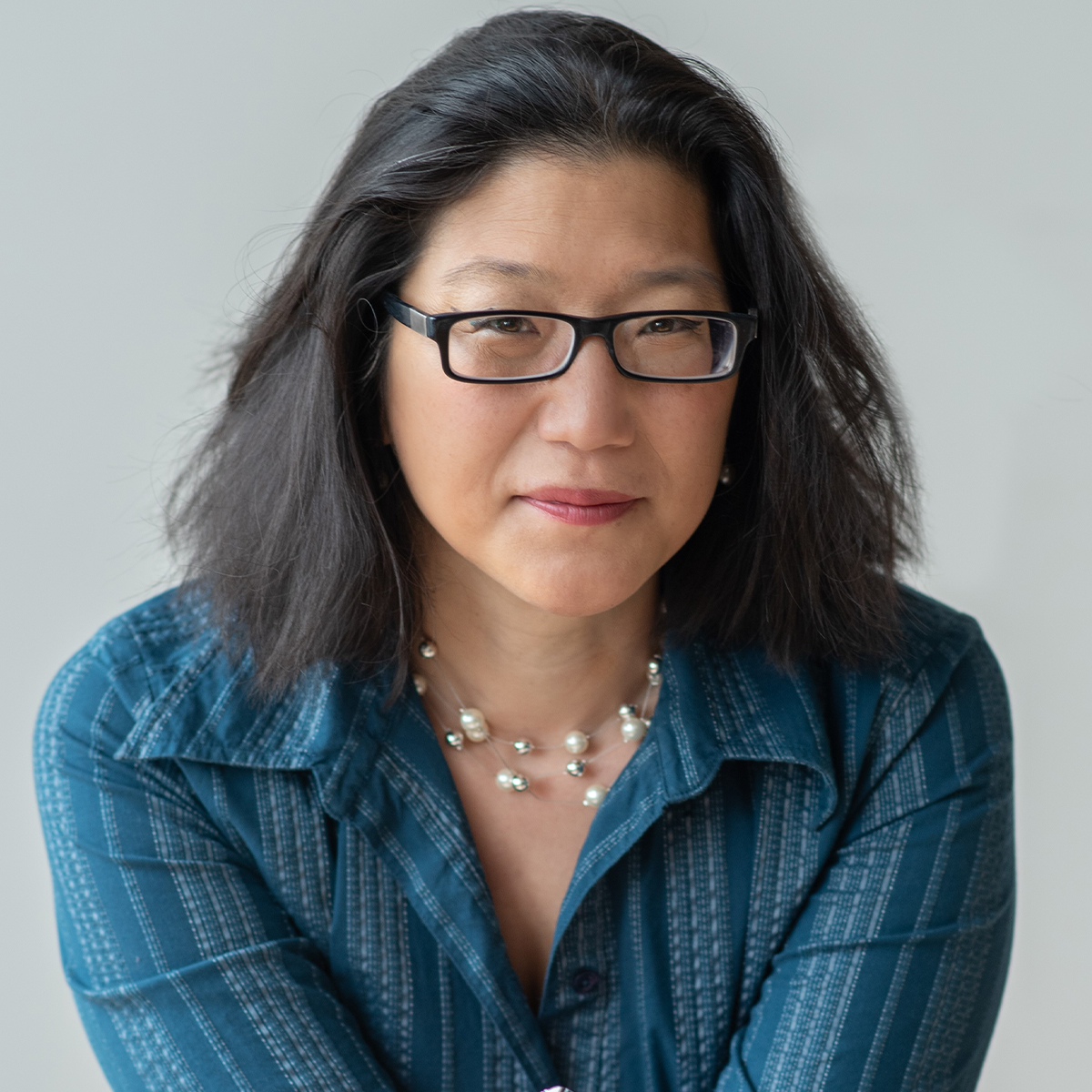 T. Susan Chang color portrait. T. Susan is a woman wearing dark-rimmed glasses, a pearl or beaded necklace, and a striped blue button-up top. She is posed in front of a neutral background.