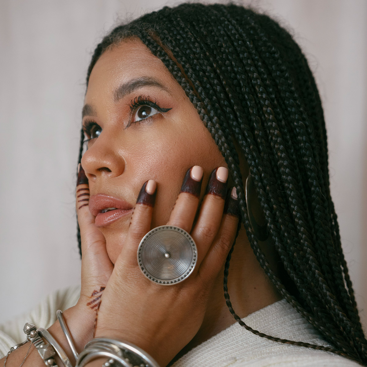 Maryam Hasnaa color portrait. Maryam is a Black woman with long, braided hair. We see her in profile and her hands are resting on either side of her face. Her eyes are cast upward and she is wearing silver rings and bracelets.