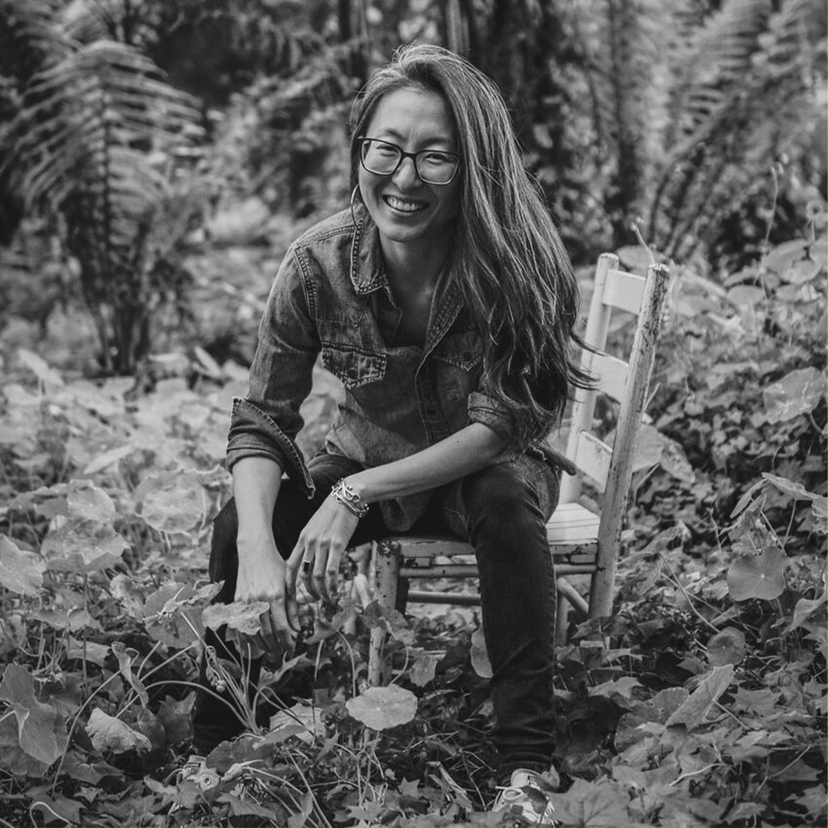 Mia Mingus black and white portrait. Mia is a Korean person smiling with long hair slung over one side of her face, large glasses and large hoop earrings. Mia is seated at the edge of a wooden kitchen-style chair in what looks like the middle of the woods.