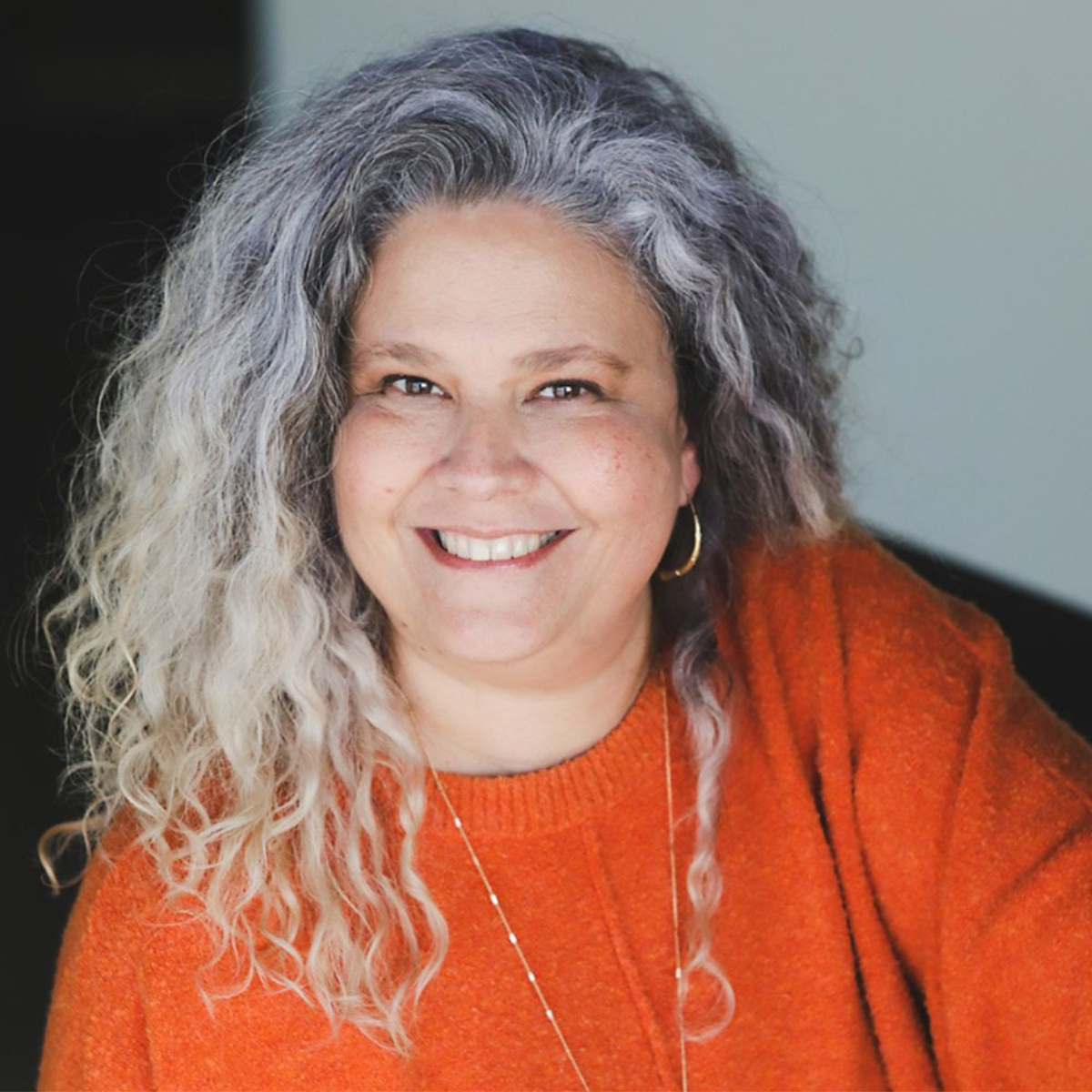 Laura Rowe color portrait. aura has silver, gray curly hair that ends past her shoulders. She is smiling and wearing a delicate, long gold necklace and hoop earrings.