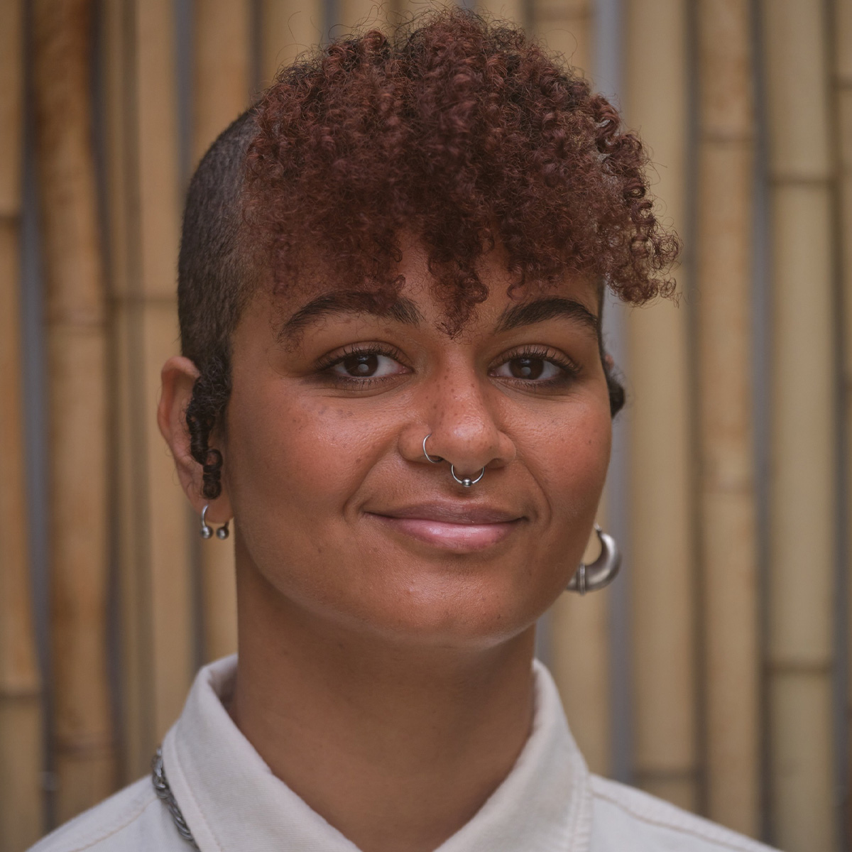 Camille Sapara Barton color portrait. Camille is a brown skinned non-binary person with their head shaved along the sides and curly brown hair create bangs over their forehead. They are smiling, wears silver earrings, and posed in front of a wall of bamboo.