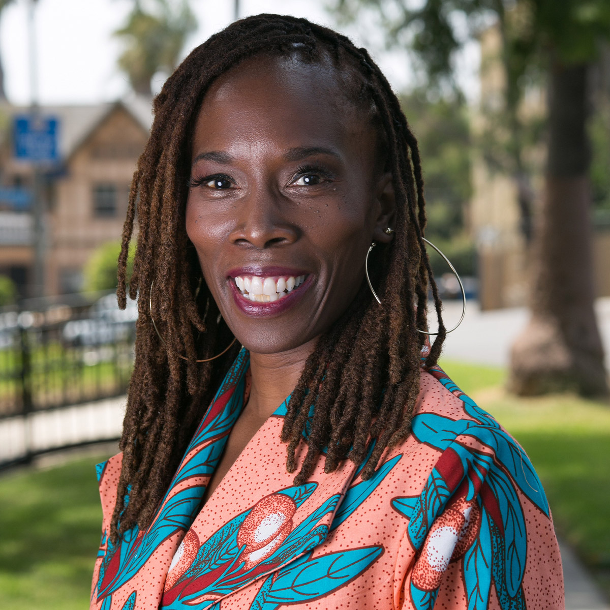 Dr. Thema Bryant color portrait. Dr. Bryant is a Black woman with dark brown to lighter brown cornrows. She is smiling and wears large thin hoop earrings and a bright pink, turquoise printed top.