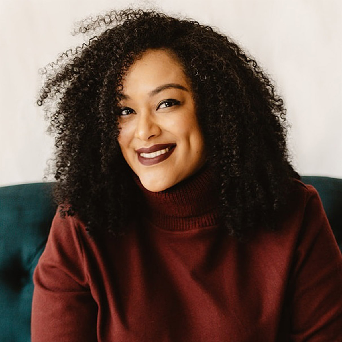 Dr. Ericka Burns color portrait. Dr. Burns is a Black woman with curly medium-length hair and is smiling, seated on an emerald-colored couch and wearing a reddish brown turtleneck.