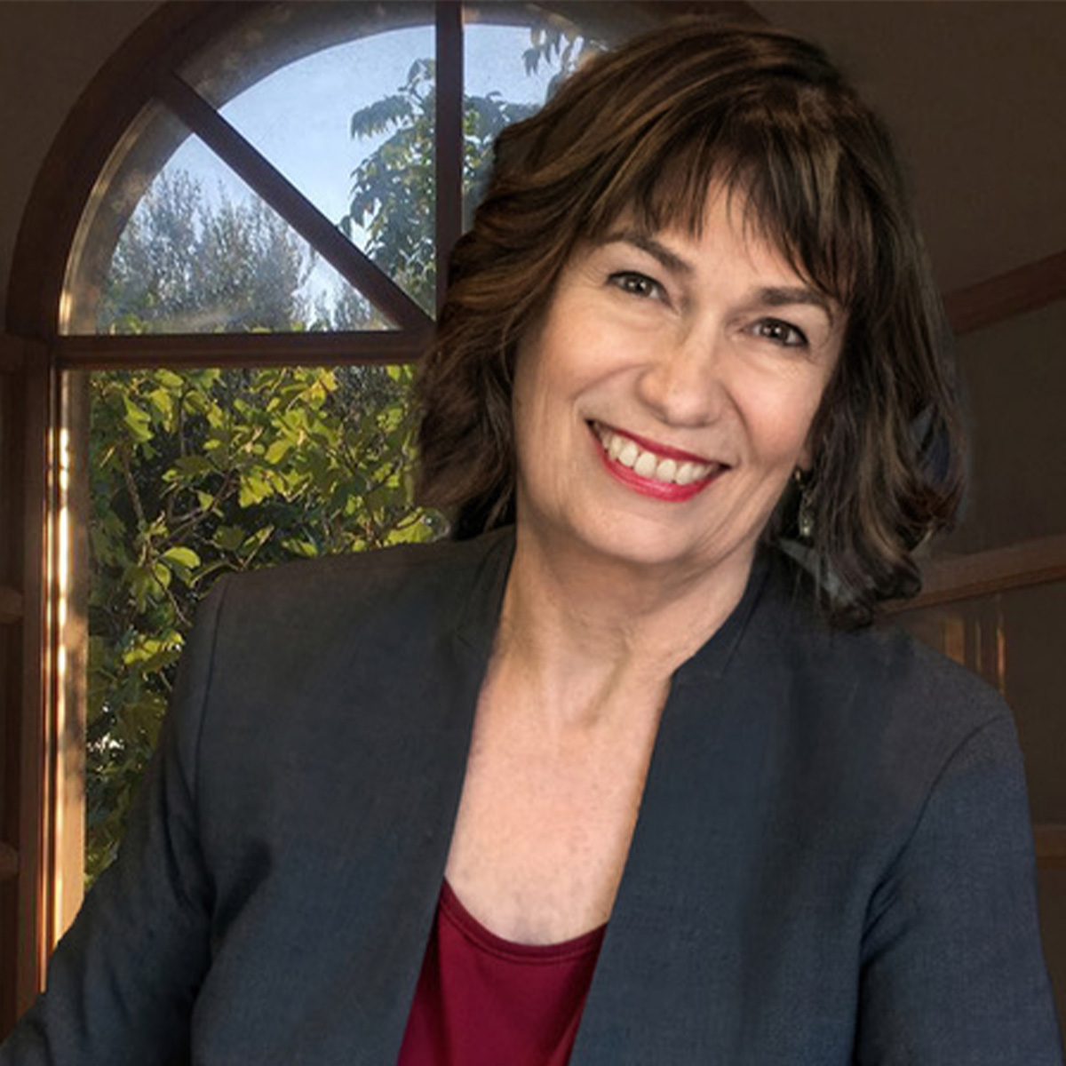 Leslie Davenport color portrait. Leslie is an older, white woman with dark brown short, wavy hair. She is smiling, wearing a dark gray-colored blazer and dark red top. She is posed in front of a large window with trees outside.