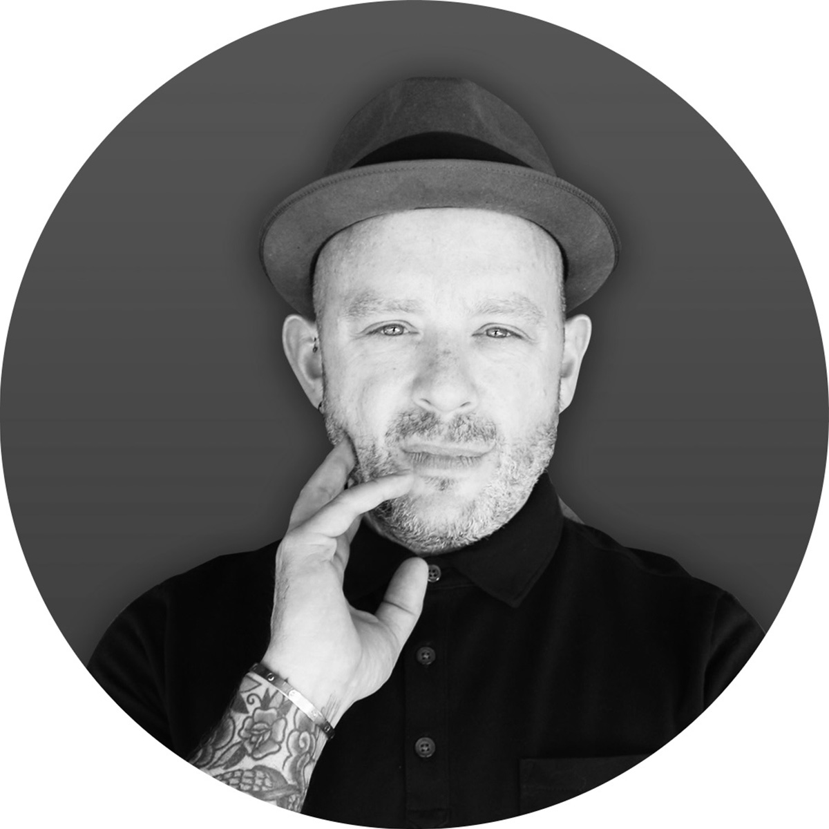 DDS Dobson-Smith black and white portrait made into the shape of a circle. DDS is a queer, nonbinary, non-disabled, middle-class, neurotypical, white British immigrant posing in front of a dark gray background, is wearing a hat slightly pushed back, has their hand resting on their cheek and chin and is wearing a dark-colored button-down.