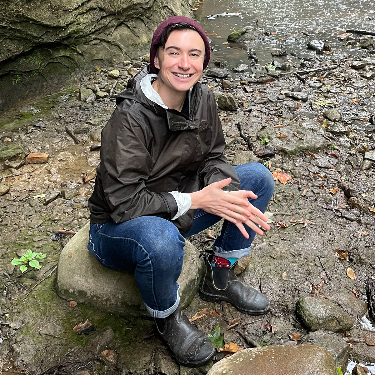 Jay is sitting on a grey green rock, surrounded by a rock formation with gray, orange and green tones. Jay's smiling face is looking towards the camera, wearing a maroon wool hat and a green raincoat with jeans. They are light-skinned, with dark eyebrows and short dark hair.