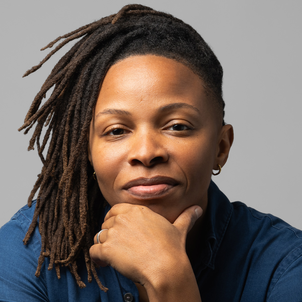Prentis Hemphill color portrait. Prentis is a Black person with medium-length locs. Their hand is resting on heir chin and they are smiling as they are posed in front of a gray background. They are wearing a blue button-down, and one small hoop can be seen.
