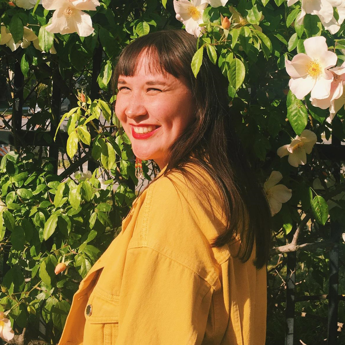 Chantal Jung color portrait. Chantal is Nunatsiavut Inuk and German and is posed outside amongst flowering trees and plants. She is turned to the left and has her head tilted towards the camera as she smiles. She has on a bright yellow coat and red lips.