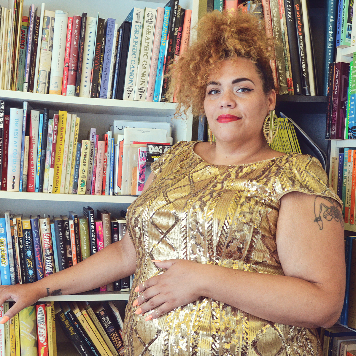 Bianca is a tall fat light skinned disabled queer AfraLatina who is in a gold dress with gold beaded geometric designs with a smile on her face and looking at the camera. Her hair is up and her blondish brown curls cascade around her head. She has on red lipstick and large round gold earrings. She stands in front of a full bookcase.
