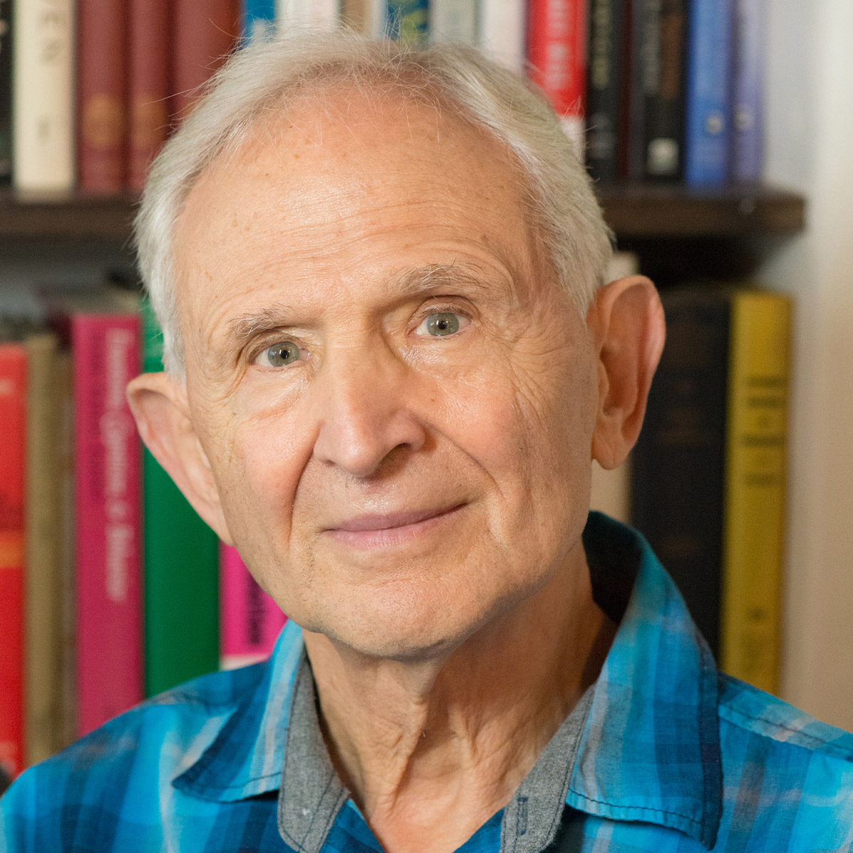 Peter Levine color portrait. Peter Levine is an older, white man posed in front of bookshelves lined with books. He is smiling and wearing a brightly-colored plaid button-down.