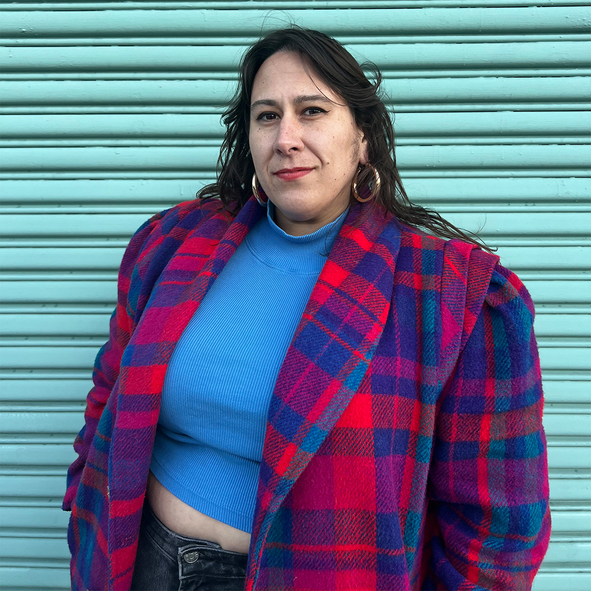 Tricia Rainwater color portrait. Tricia is a Choctaw woman standing in front of a bright teal curragated wall. She is smiling and is wearing a plaid coat, a blue mockneck cropped top, and jeans. She has straight, medium-length hair and is wearing hoop earrings.