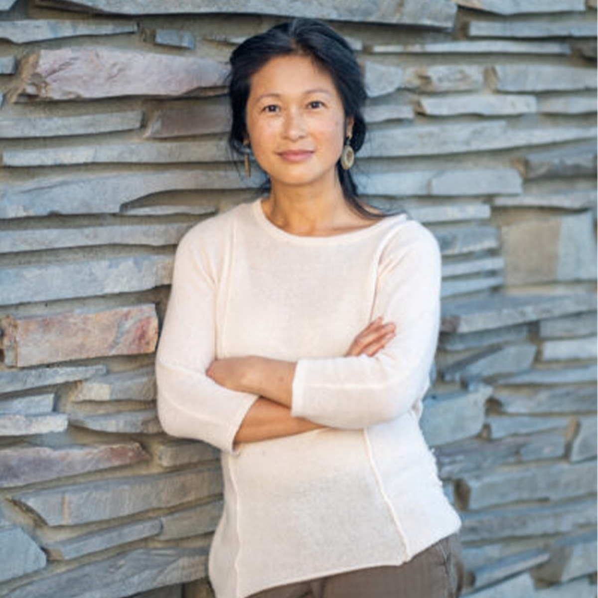 Linda Thai color portrait. Linda is a Vietnamese woman who is standing and leaning against a stone wall. Her arms are crossed in front of her and is smiling.