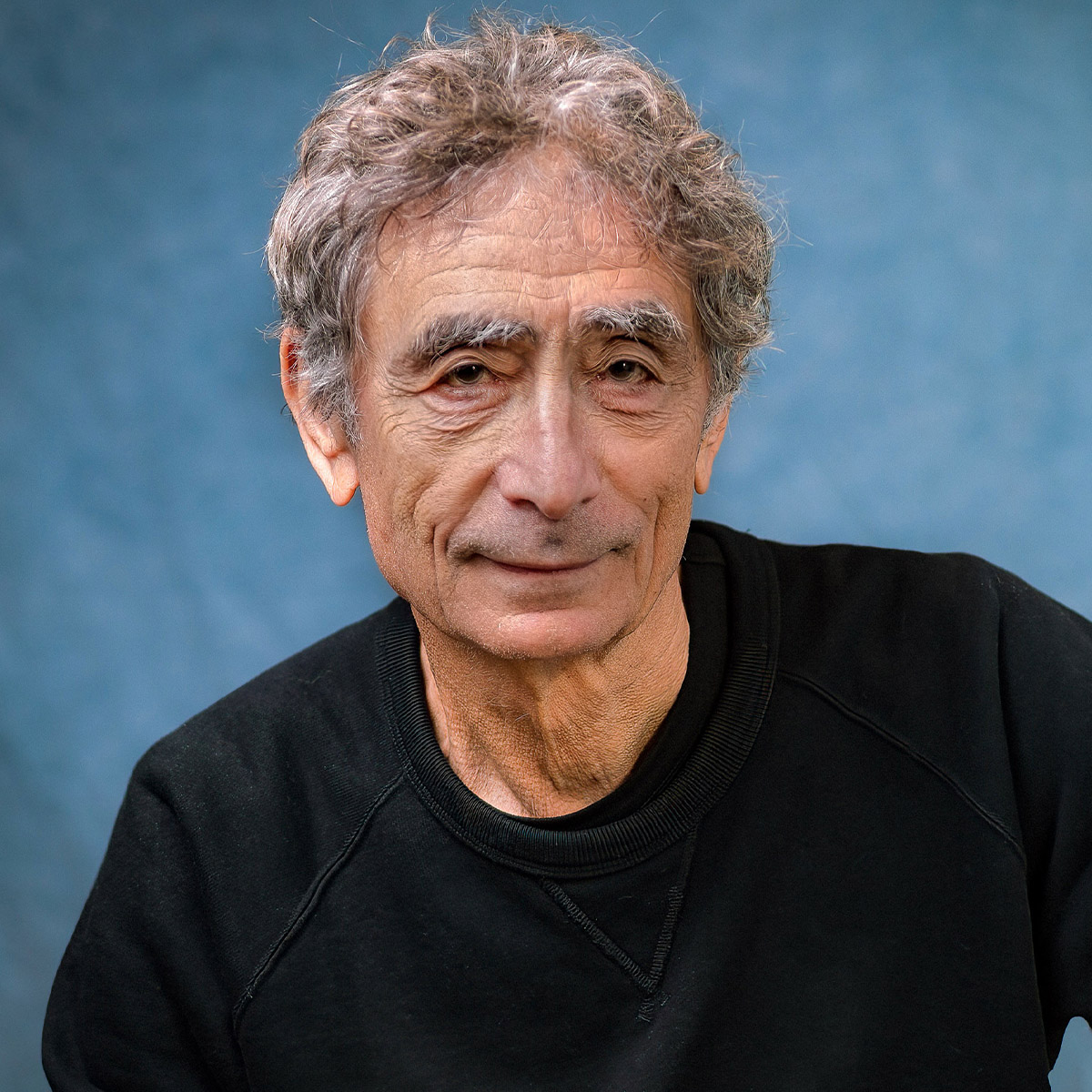 Dr. Gabor Maté color portrait. Dr. Maté is a Canadian-Hungarian older white man who is posed in front of a soft blue background and is wearing a dark-colored sweatshirt.